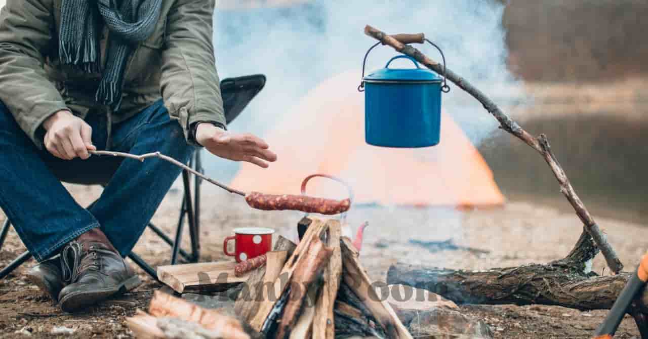 焚き火でソーセージを焼く人と吊るされたケトル、安全に配慮したキャンプ調理の様子