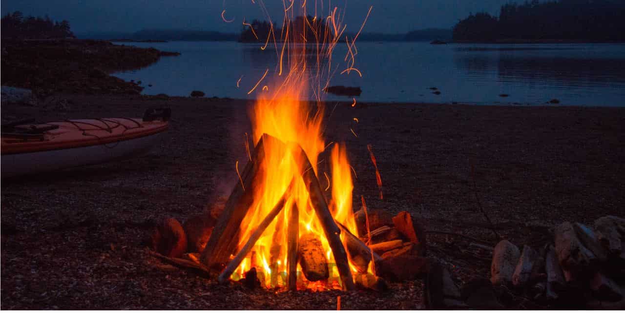 夕暮れの湖畔に燃え上がる焚き火。静かな夜の自然と向き合いながら、虫や音との付き合い方を考える場面。
